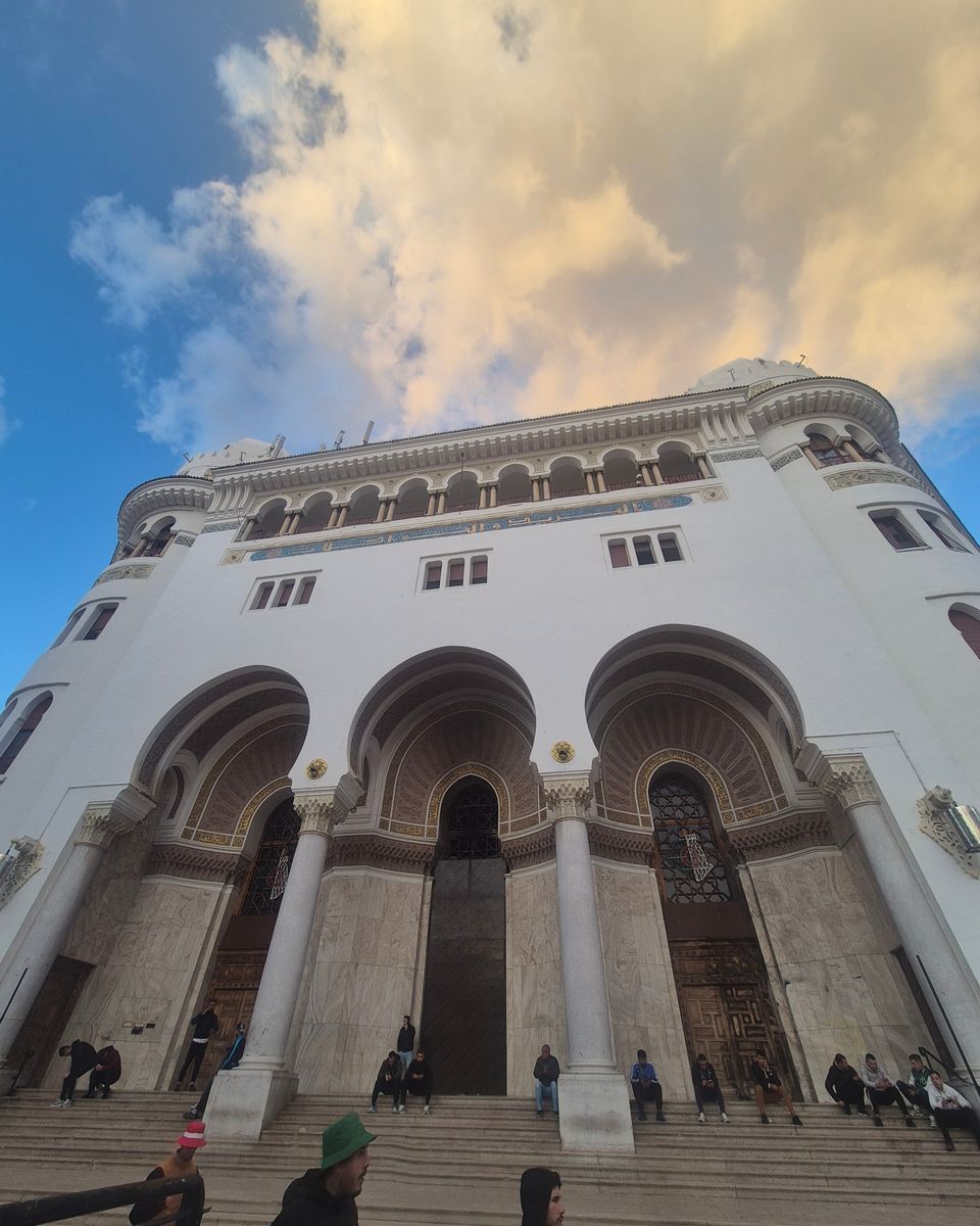La façade blanche de la Grande Poste d'Alger, avec ses arcs de style néo-mauresque, sous un ciel bleu et nuageux