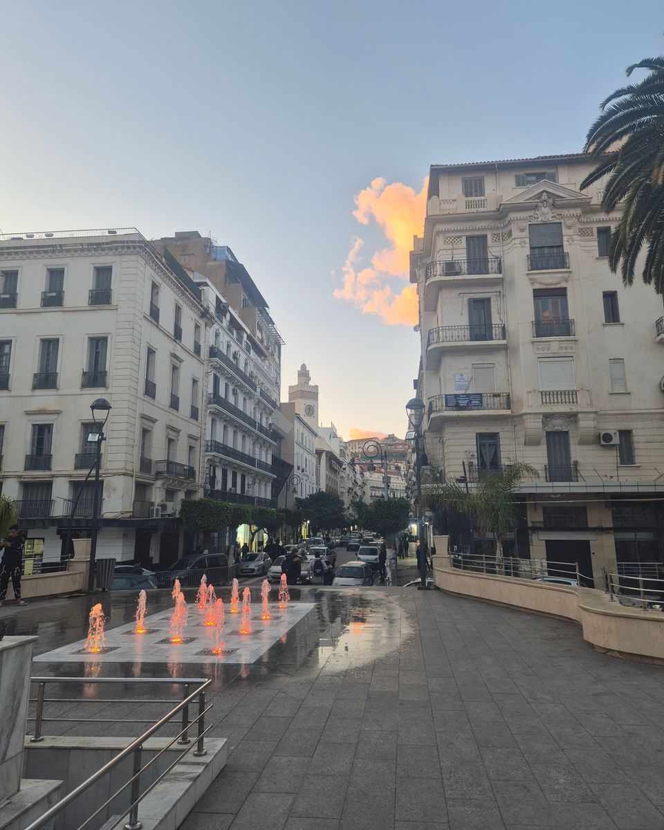 Une place piétonne d'Alger avec des jets d'eau et des immeubles blancs, au coucher du soleil