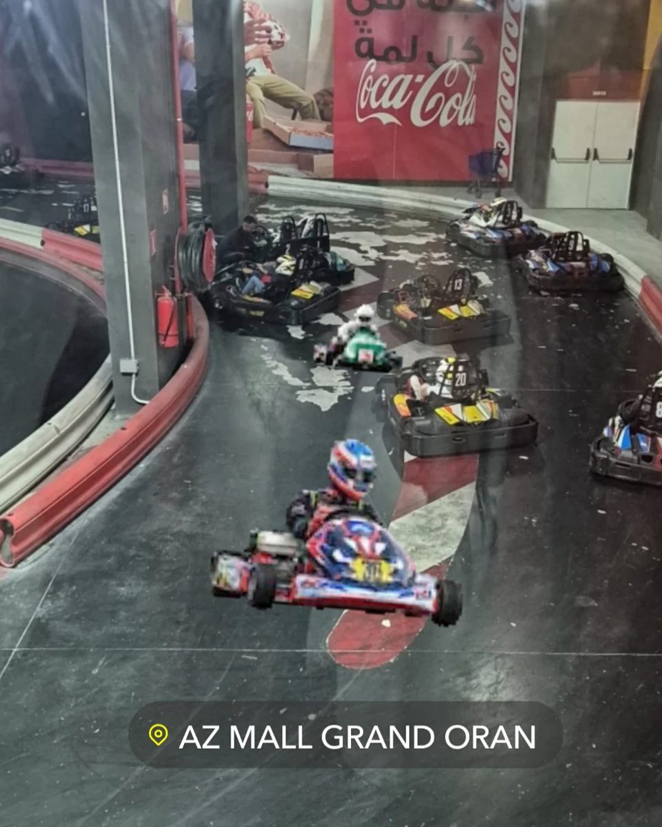 Une piste de karting en intérieur, avec plusieurs karts et des pilotes casqués, au centre commercial AZ Mall Grand Oran
