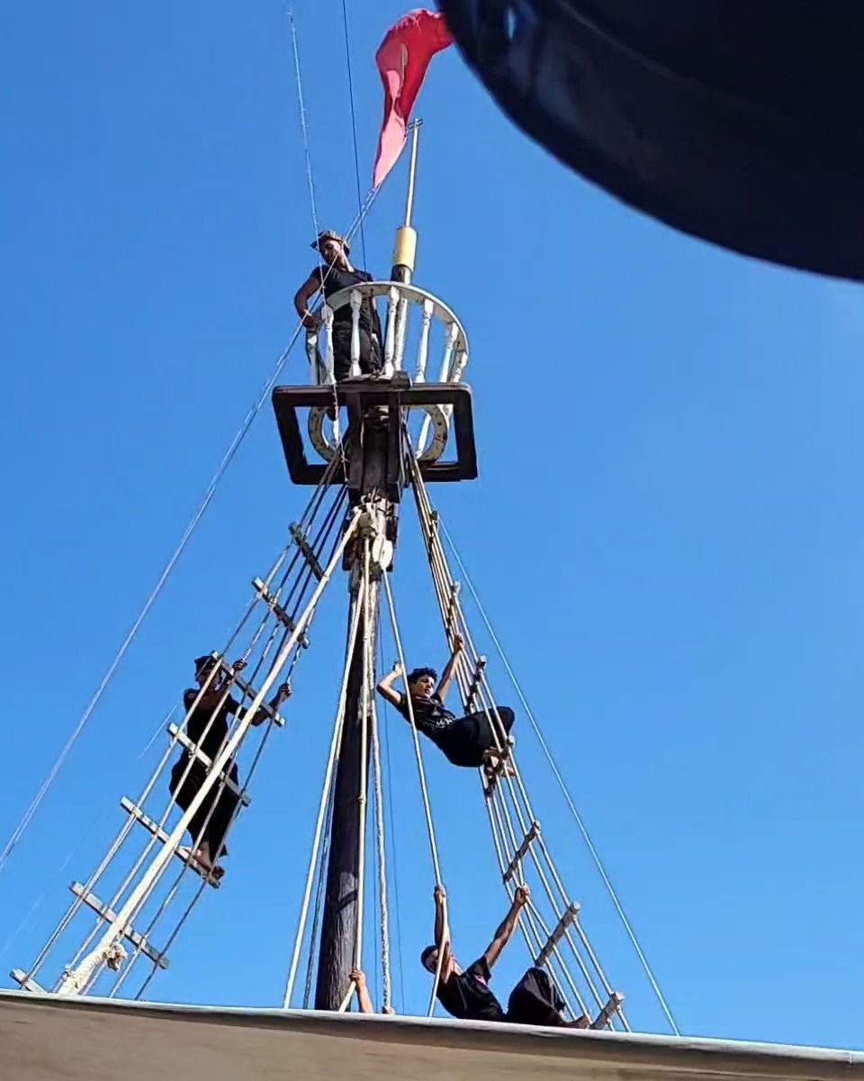 Le mât d'un bateau de croisière décoré en bateau pirate, avec un drapeau rouge et des passagers qui grimpent aux cordages