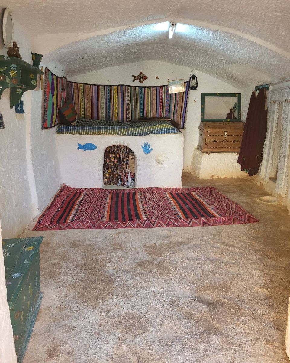 Une chambre taillée dans la roche blanche, avec un lit maçonné, un tapis rouge et un coffre en bois