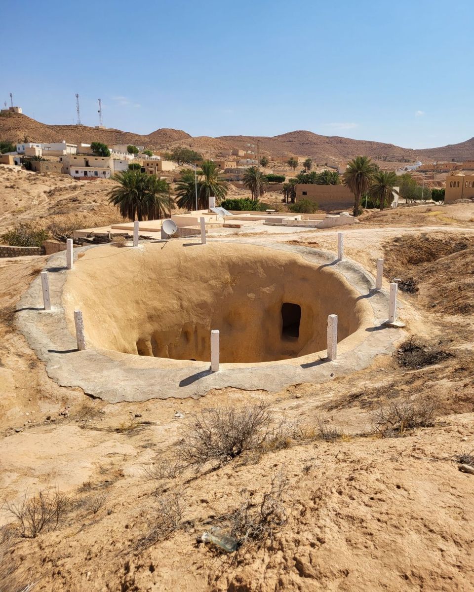 Une maison troglodyte vue d'en haut : un grand trou rond creusé dans le sol, avec une porte taillée dans la paroi, et le village de Matmata derrière