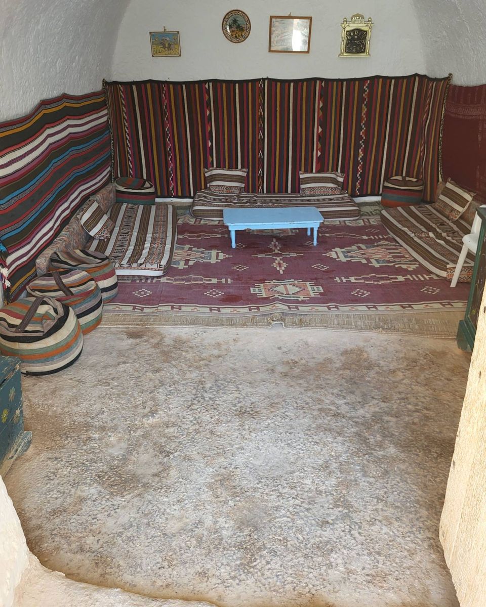 Le salon d'une maison troglodyte, aux murs couverts de tissages rayés de couleur, avec des matelas au sol et une petite table bleue